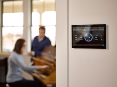 A hallway in the foreground showing a wall panel screen with a Control4 interface, as a couple is at the piano in the background.